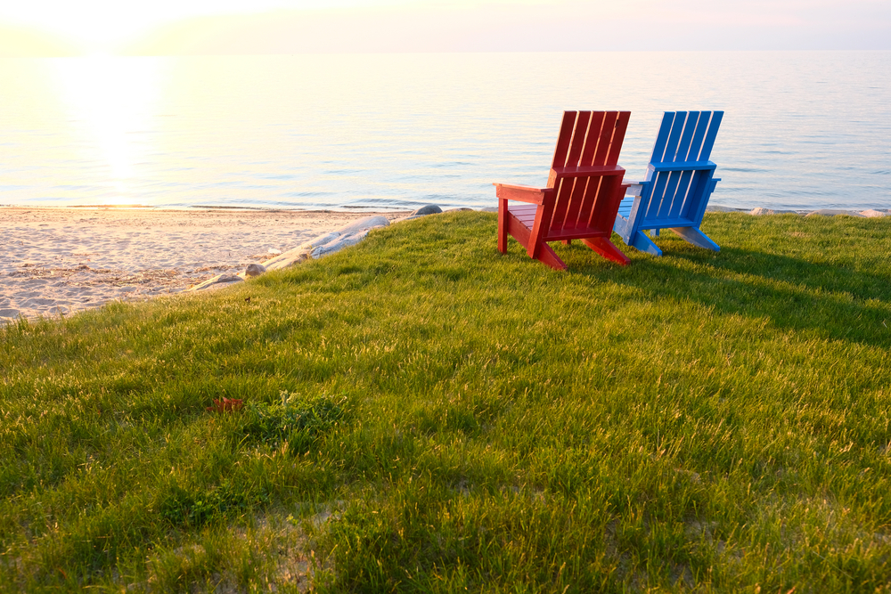 Sandy Michigan Beach and gently lapping waves as sun sets with red and blue Adirondack chairs in foreground.