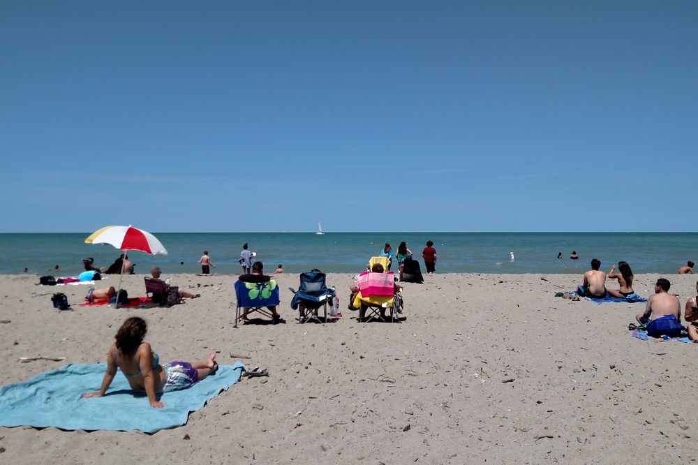 Sandy Ohio beach with people sitting in sun, Lake Erie waters and tiny sailboat on horizon.