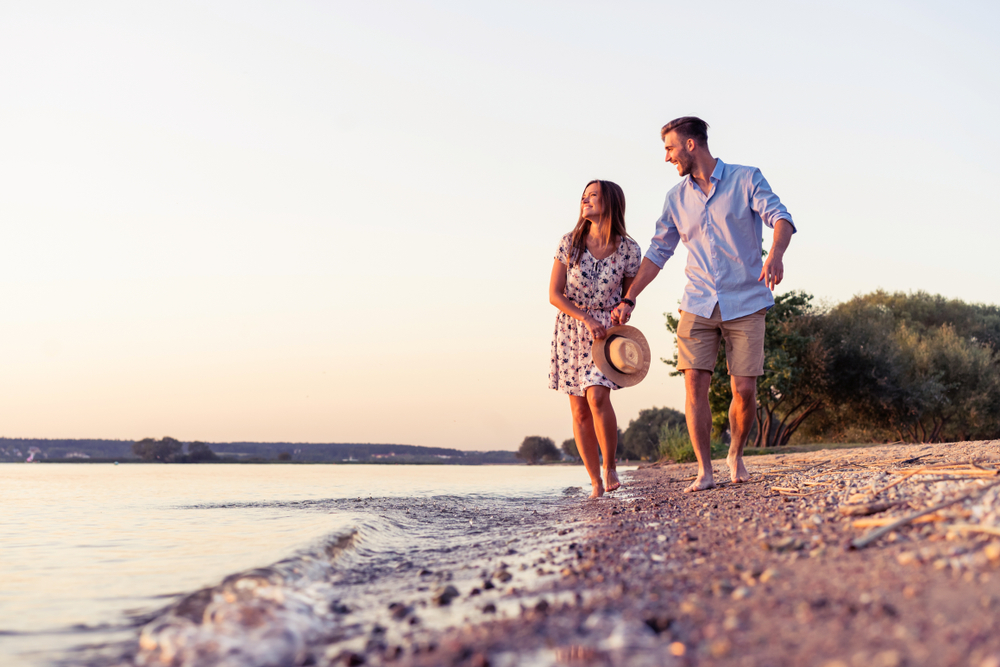 Smiling couple walking on rocky beach with water on left and trees in background.