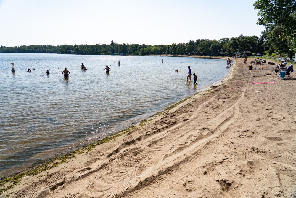 Sandy beach with people swimming in lake with sandy beach in foreground.