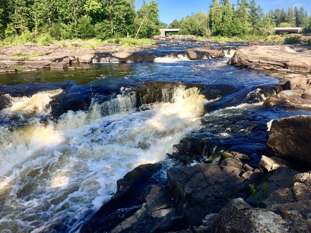 Small Minnesota waterfall from river above it flowing rapidly over rocky bottom.