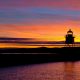 Sky lit at sunset with pink and yellow with lighthouse silhouetted in background with still water in foreground of Grand Marais MN