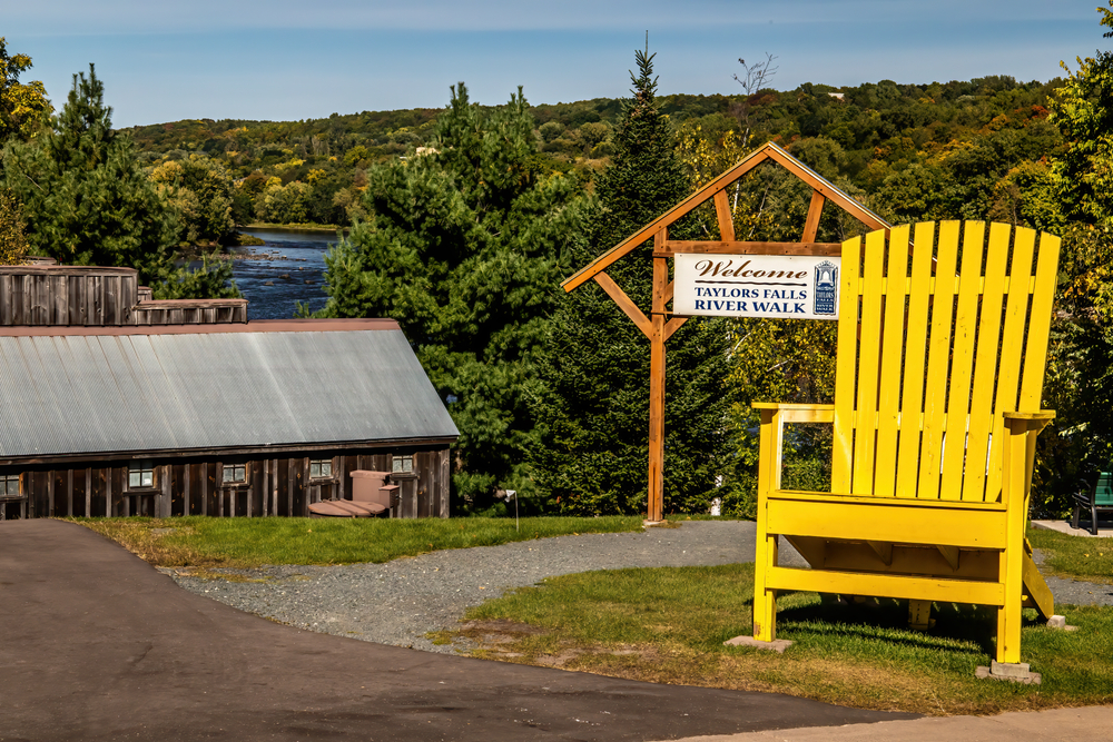 Large yellow Adirondack chair near welcome sign leading to Taylors Falls Riverwalk with water in background