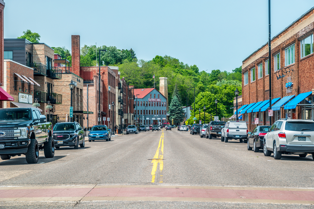 Main street with quaint building on either side with cars parking along sides. Green trees in background.
