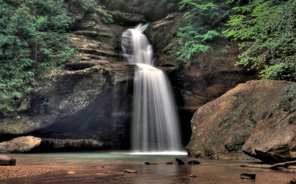 Cascading waterfall through rocks into small green pool of water. Trees surrounding on each side.