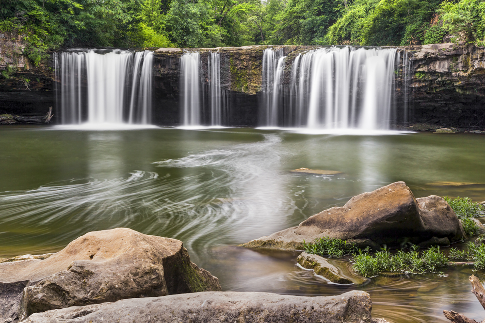 Water pours over rock ledge into large pool of water below. 