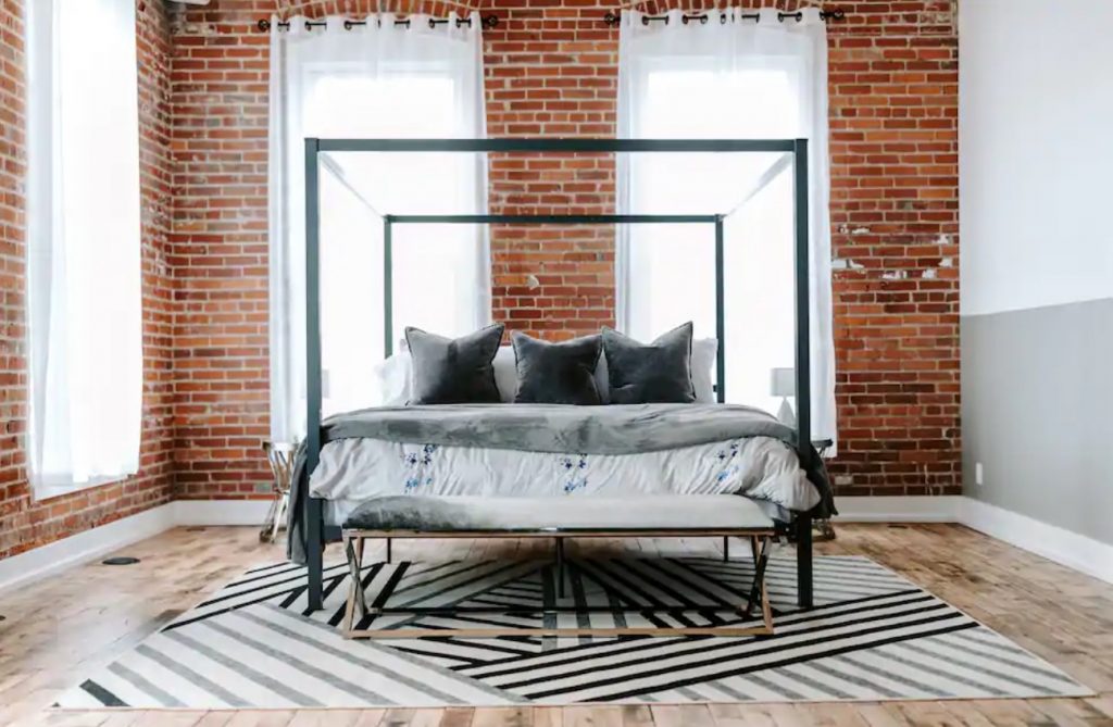 A simple but beautifully decorated king size bed in a space with exposed brick walls and floor to ceiling windows