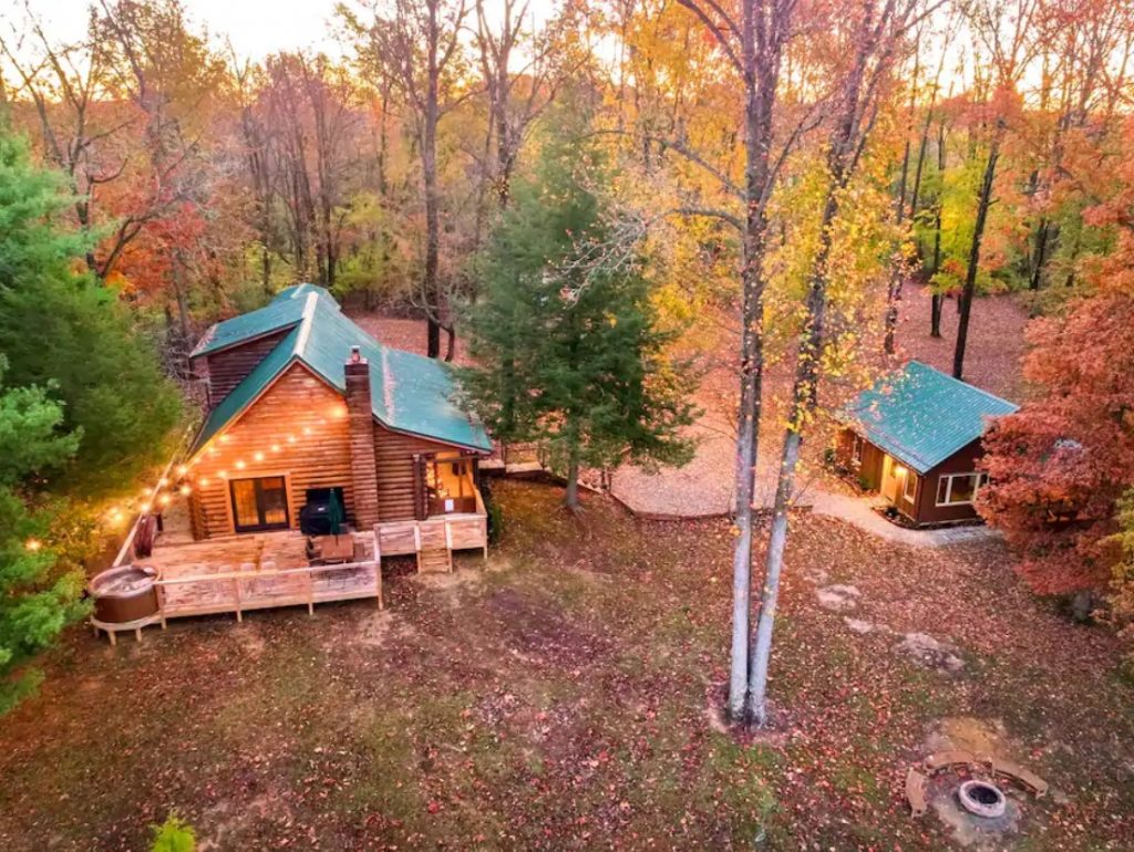 An aerial view of a large log cabin with string lights on a deck and a smaller log cabin in the woods surrounded by trees in the fall one of the best Airbnbs in the midwest