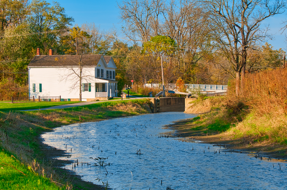 White homes in front of walking trail with river in foreground and autumn trees 