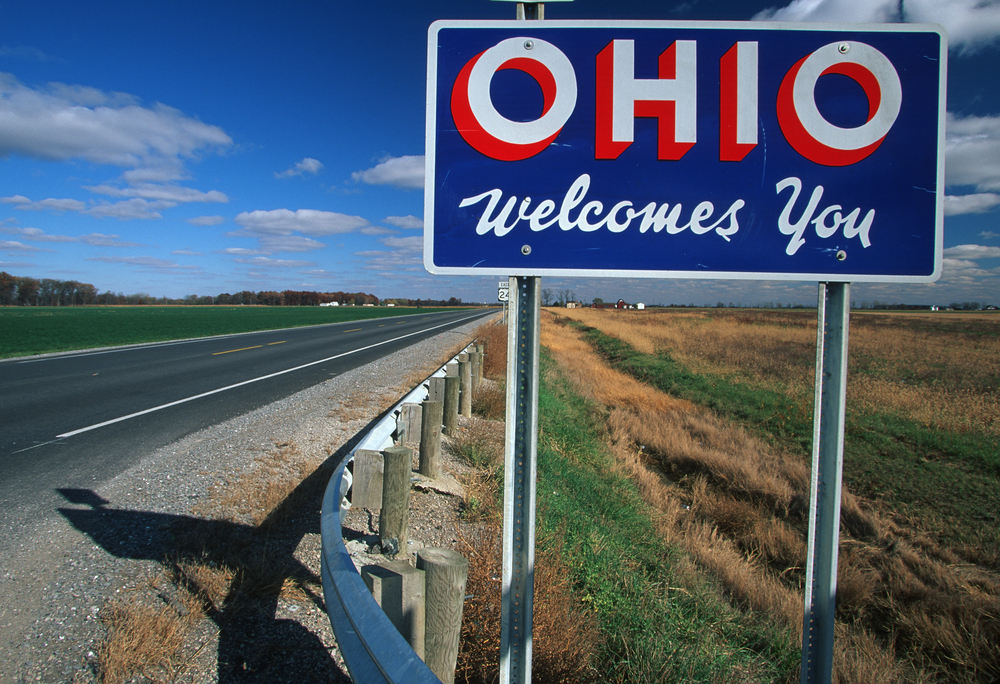 Bright blue Ohio Welcomes You road sign alongside highway with farmland on either side.