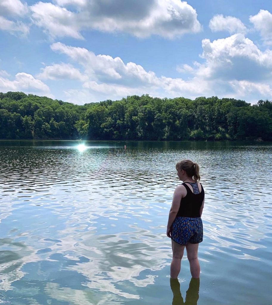 Girl stands looking out over sunny lake lined with lush, green trees at park.