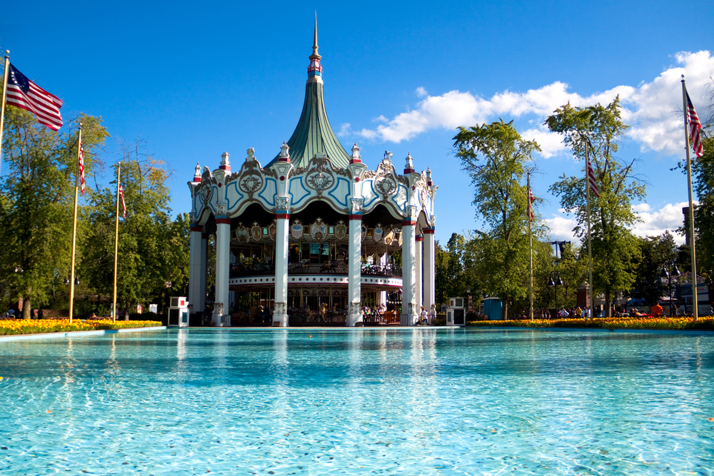 A merry-go-round by a large blue lake surrounded by flowers and American flags blowing in the wind