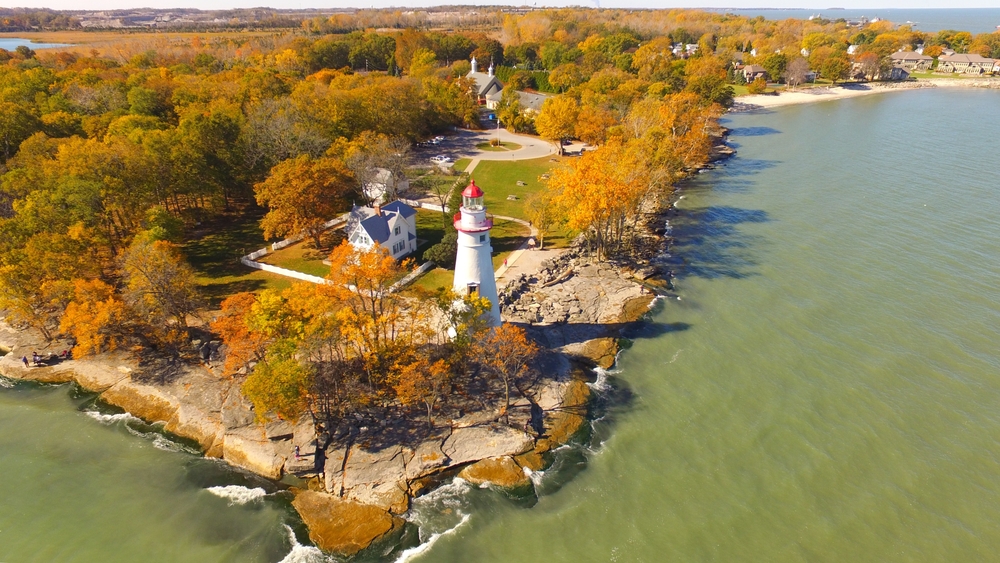 Aerial photo of white lighthouse with fall foliage in Ohio surrounding it with Lake Erie waters  in foreground.