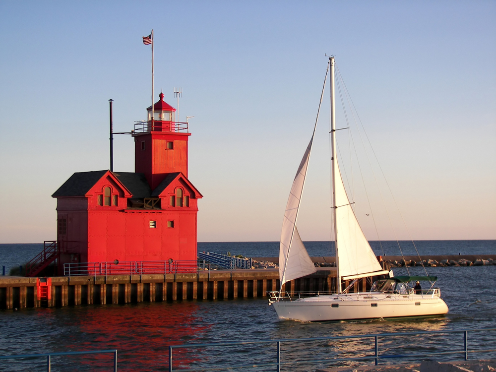 A large red Michigan lighthouse on the end of a small pier with a sail boat in front of it 