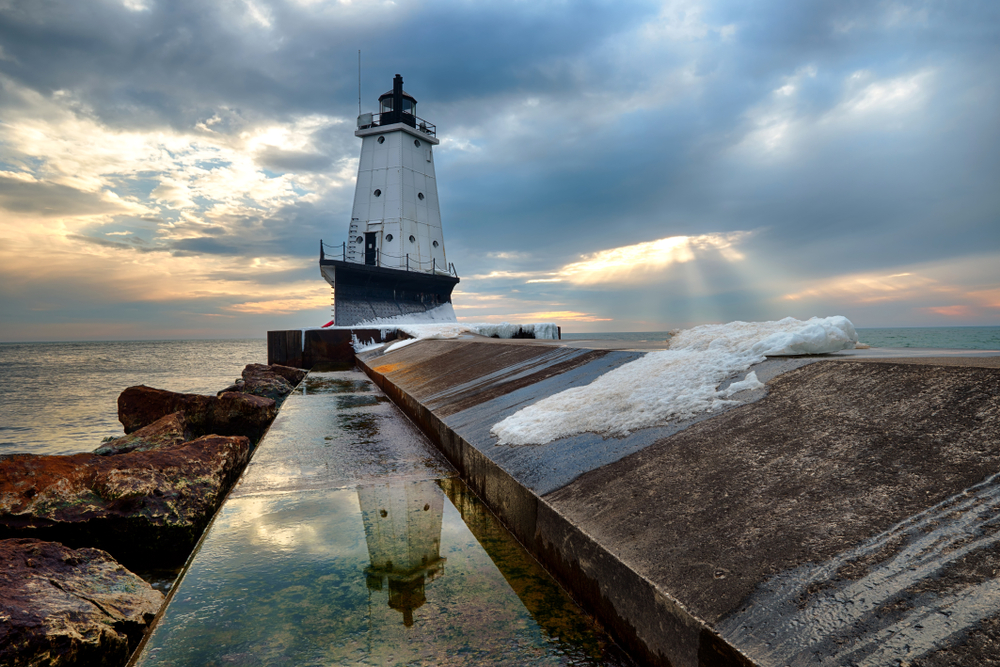 A tall white metal Michigan lighthouse on the end of a long pier with  large rocks to left and sunset in background.