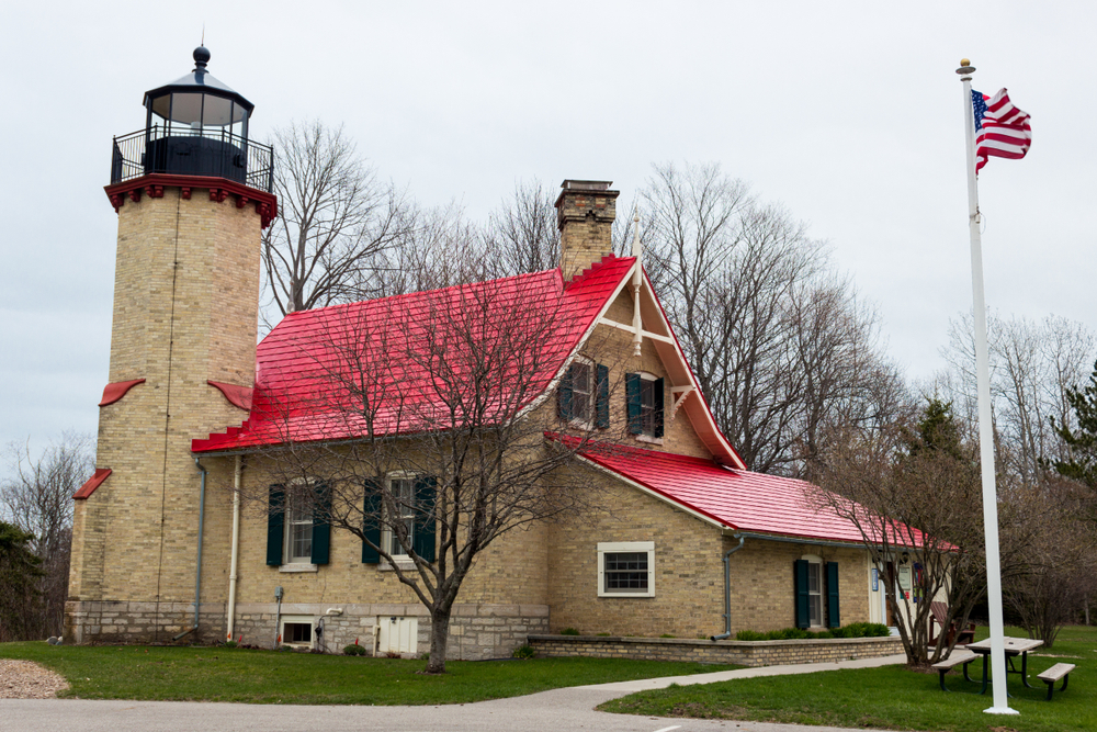 A tan brick lighthouse with a red roof and surrounded by a green lawn and trees with American flag blowing. Michigan lighthouse.