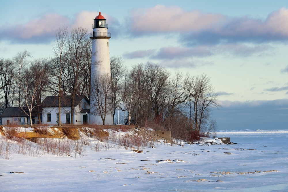 A tall white lighthouse in Michigan on the edge of lake in winter with snow all around.