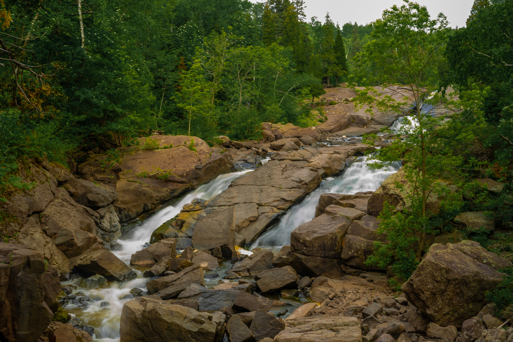 Two small waterfalls that run through large splits in a large rock base on the side of a mountain surrounded by trees