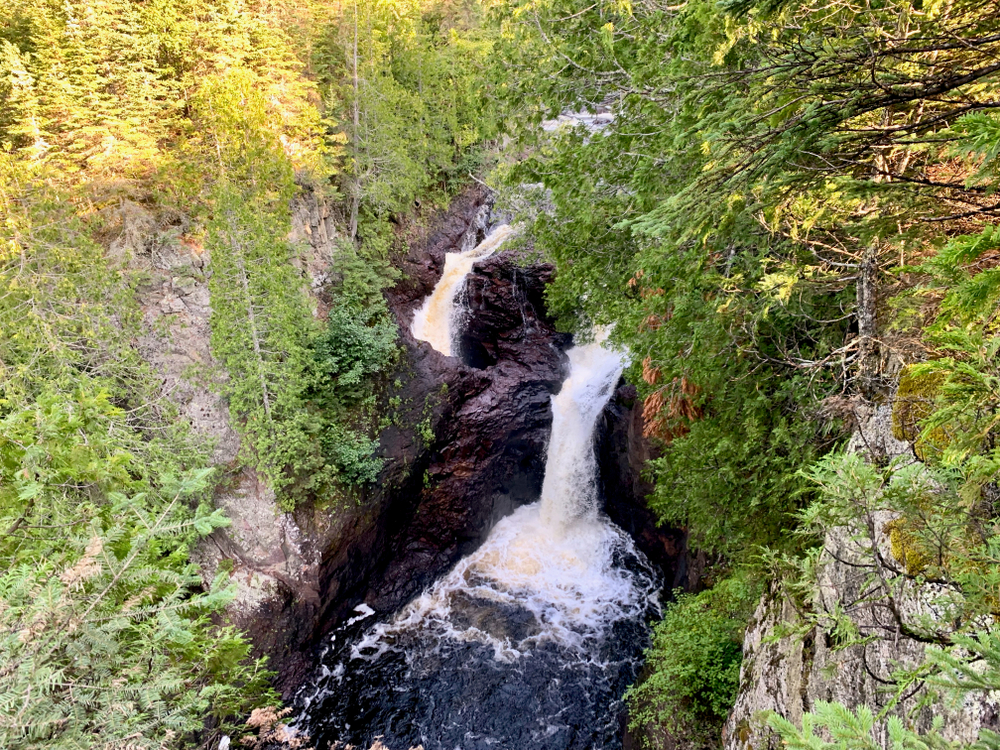 Two large waterfalls surrounded by rocky cliffs and large trees where one runs into the river while one waterfall appears to disappear underneath the rocks.