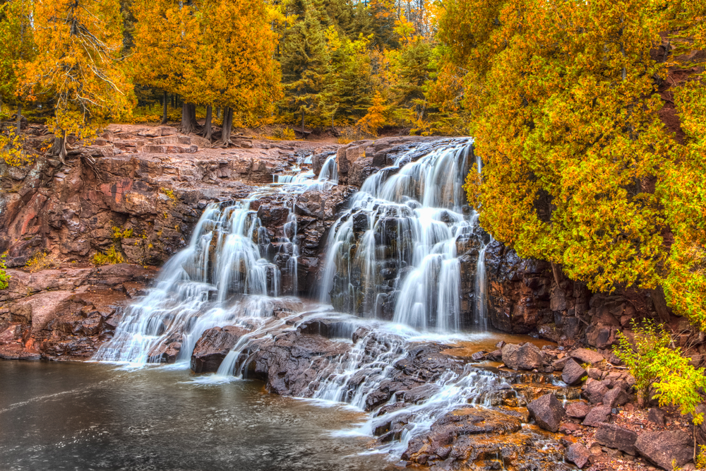 A large rushing waterfall over a rocky cliffside surrounded by yellow trees in the fall waterfalls in minnesota