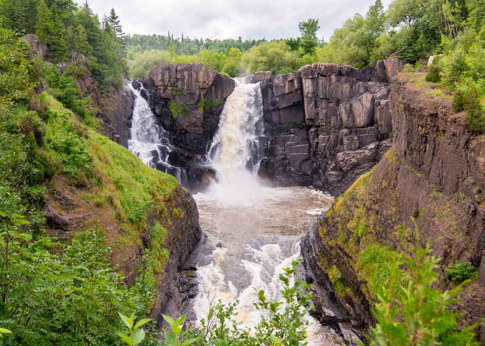 Two large waterfalls running over a rocky cliffside that runs into a river surrounded by moss and lush greenery waterfalls in Minnesota