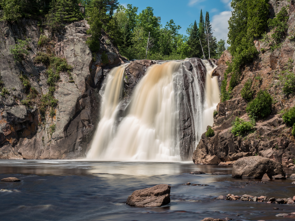 A large rushing waterfall on a sunny day surrounded by rocky cliffs and tall green trees that runs into a pool of water