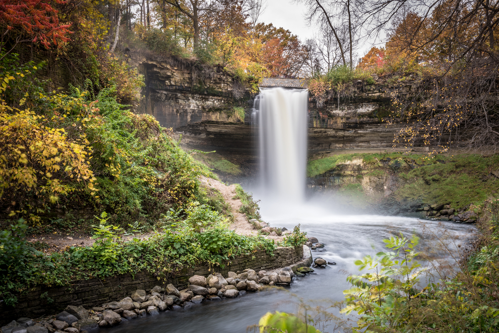 A large waterfall on the side of a rocky cliff that runs into a river surrounded by greenery