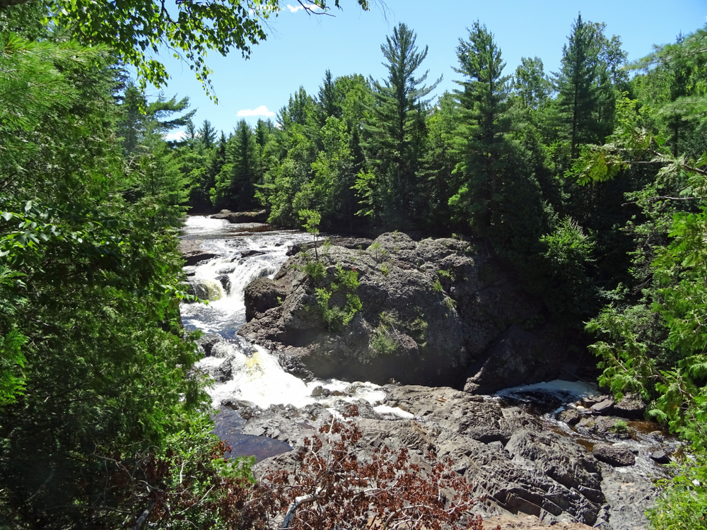 A portion of the Potato River Falls on a sunny day with blue skies surrounded by large green trees and interesting rock formations