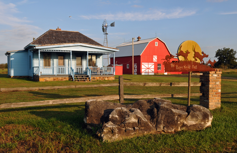 Two buildings restored including a blue farm home and a classic red barn with a windmill in the replica town of Red Oak II in Missouri on a sunny day