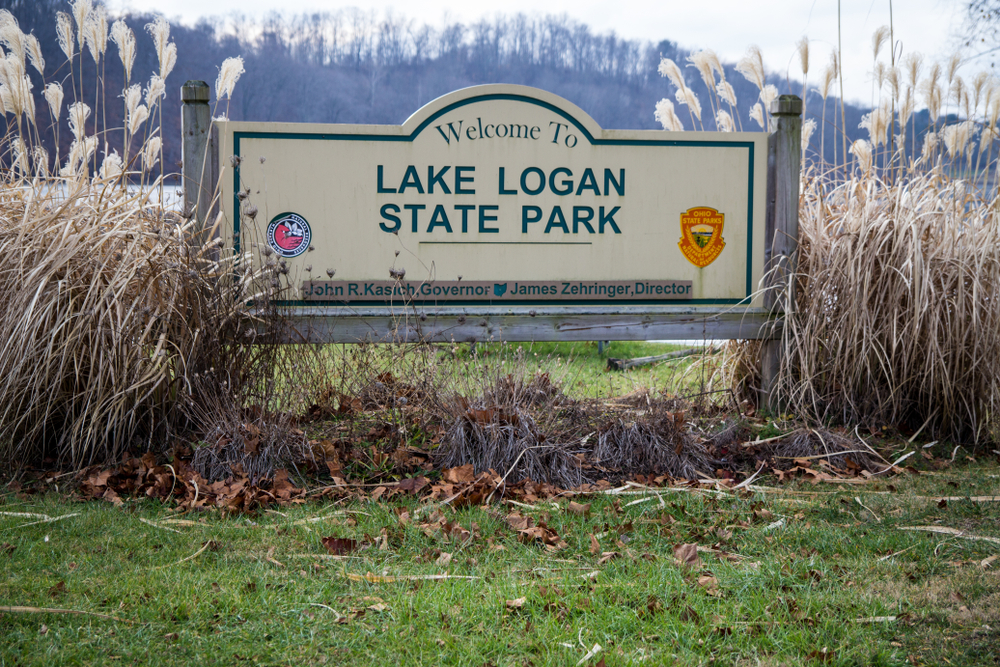 Tan welcome sign with green letters "Welcome to Lake Logan State Park' surrounded by dried brush.
