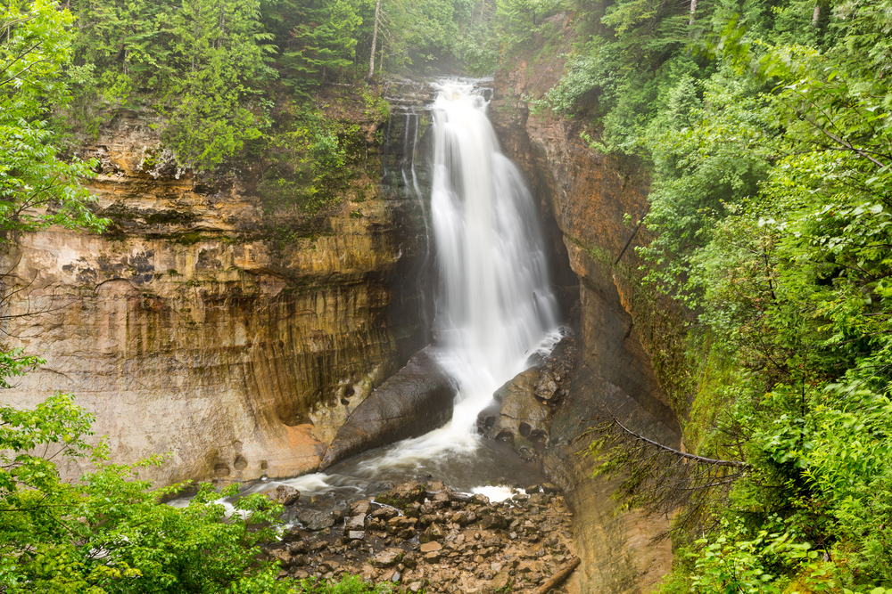 A large waterfall tumbling off a cliff with green foliage on and around the cliff. One of the waterfalls in Michigan