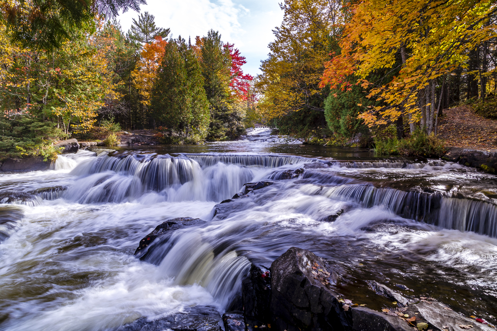 Water tumbling on rocks with a waterfall and fall foilage in the background in an article about waterfalls in Michigan