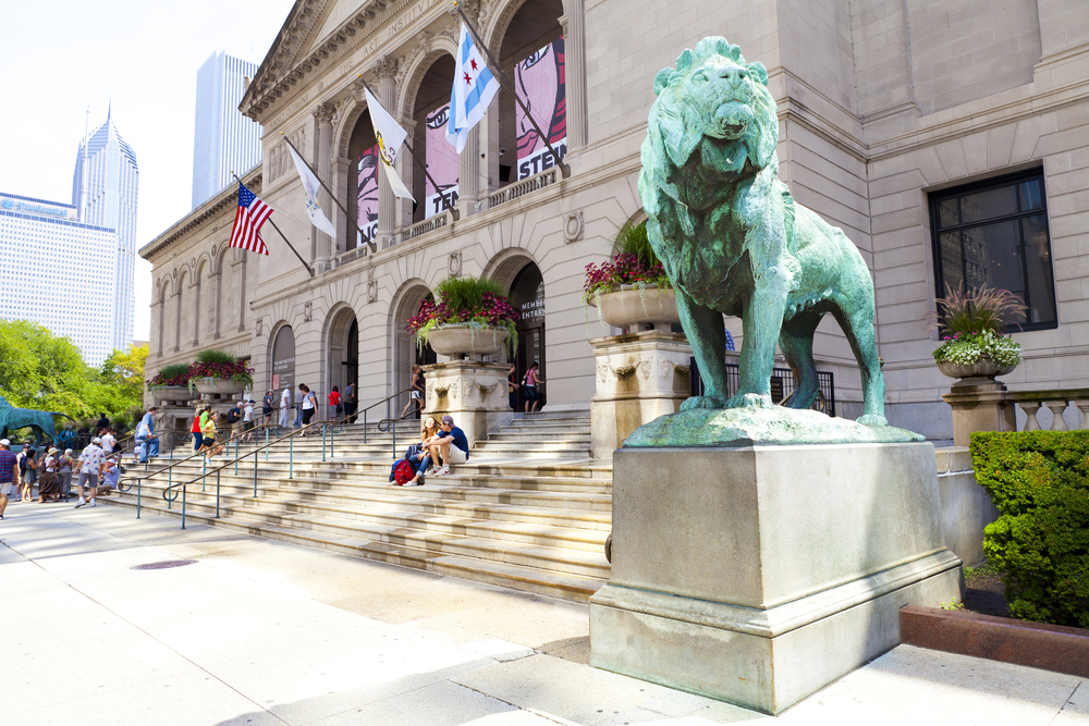 Historic light-colored building with decorative arches and flags flying with green statue of animal in foreground.