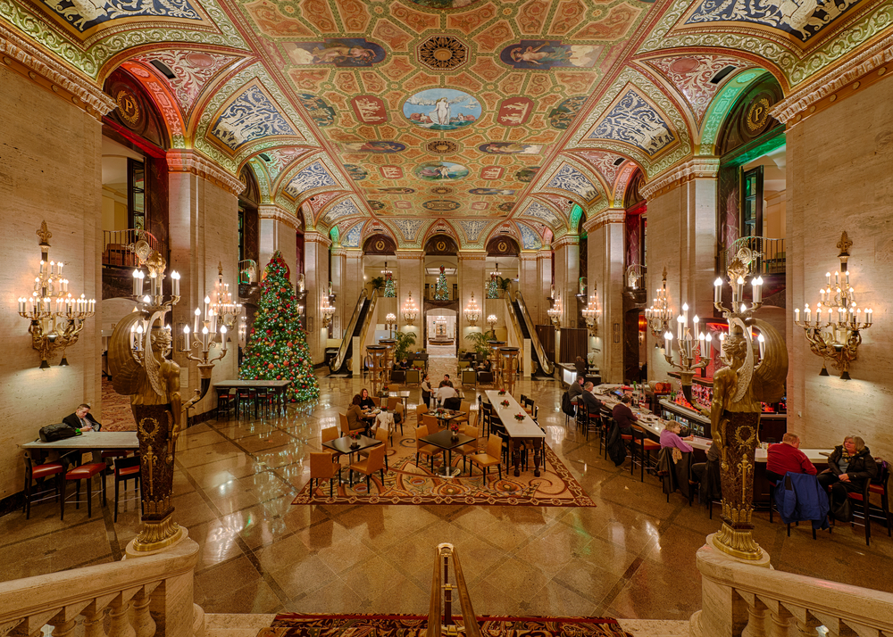 Opulent hotel lobby with chandeliers on walls, fancy colorful ceiling and decorated Christmas tree on left.