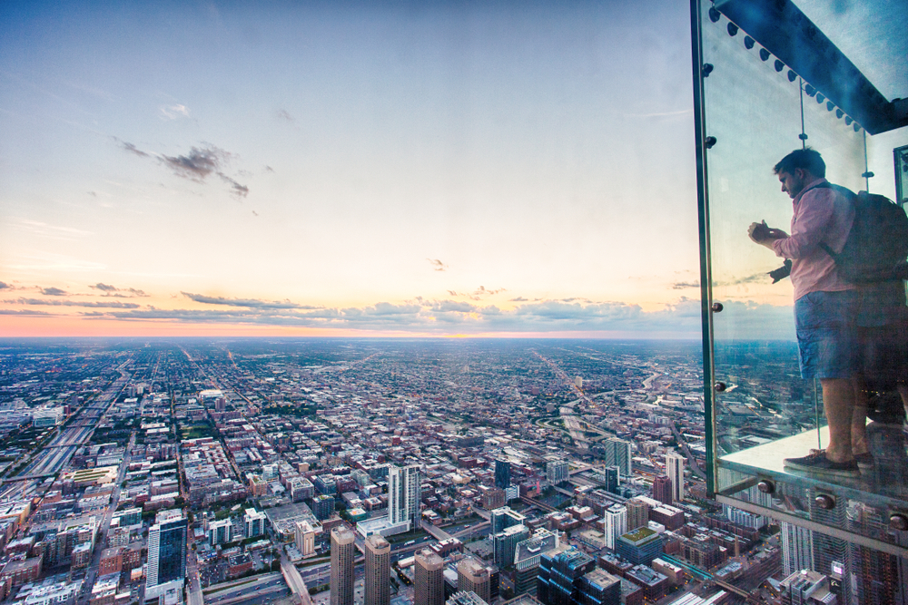 Glass box extending out with male visitor holding camera. City skyline in background.