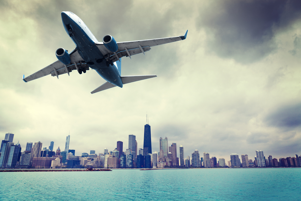 Airplane flying over blue waters with city skyline in background.