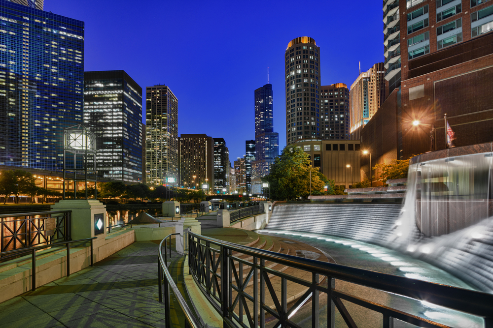 Concrete path in city at night with skyscrapers all around it lit brightly.