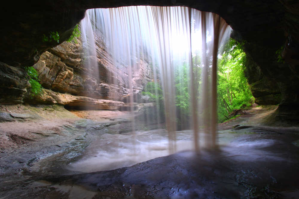 behind view of waterfall flowing  into rough waters below, with sandstone walls and green trees in background.