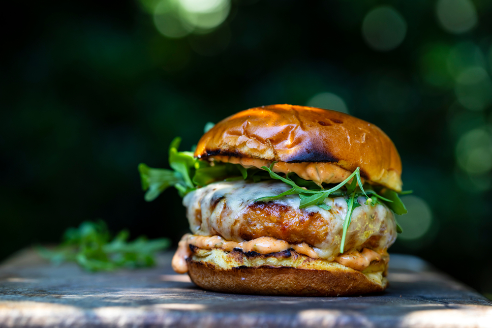 A charizo burger with a creamy orange sauce, cheese, and spinach. The bun is toasted. The burger is sitting on a dark material and there is a green and black bokeh background. 