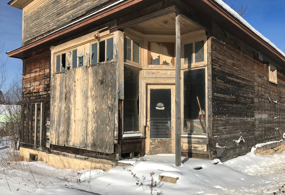 An abandoned building in a mining town in Michigan. It looks like it was once a store front. It is badly decaying with wooden sides that looks like they have mold and mildew on them. One side of the windows are boarded up, but the top  windows are broken. There are two large windows and a glass door in that make a corner in the front of the building. Those windows are still mostly intact. The old trim on the windows and doors is a faded and dirty yellow. There is snow on the ground in front of the dilapidated building. 