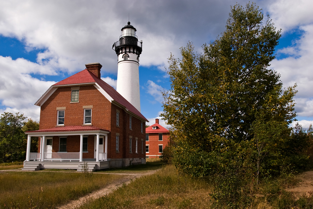 The Au Sable Light station. It is a brick building with a small white lighthouse behind it. The house has a red roof, a small front porch, and white trim. There is another smaller brick building behind it. In front there is grass and trees.