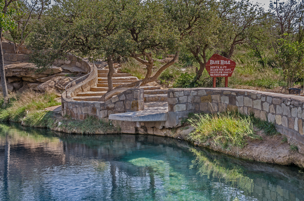 A crystal blue lake with grass and shrubs around it. There is a stone wall along the side and stone steps that lead down to the lake. There is also a sign that says "Blue Hole" and lists different information about the lake. 