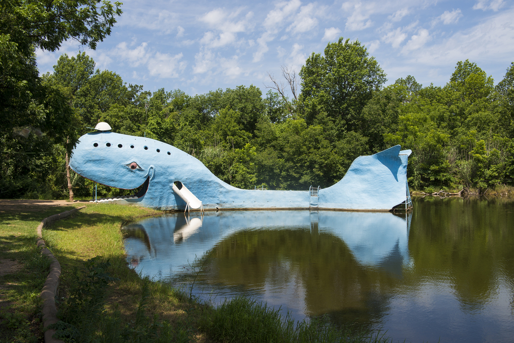 A large metal blue whale floating on the shores of a lake. On the side of the whale, a white slide comes out and there is also a ladder. The whale's mouth is open on the shore of the lake. There is grass and trees all around the lake. Its one of the best route 66 attractions