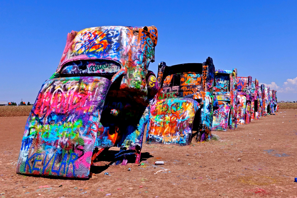 Several Cadillac's buried in the dirt about half way. They have been spray painted with graffiti in tons of different colors. It is a bright sunny day.