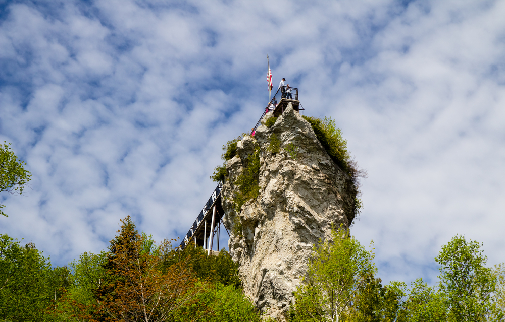 Looking up at Castle Rock, which is a unique rock formation on the side of a hill. It is a tall rock with a bridge to it. There are a bunch of trees under the rock and it is a sunny day with a kind of cloudy sky.