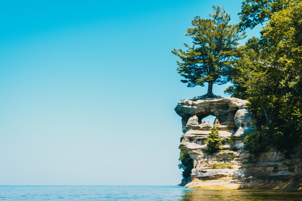 the view from the shore of the lake of Chapel Rock. It is a rock formation that goes into the lake and has a hole in the top center of the side of the rock formation. There is a tree growing directly on top of the rock formation, and trees surrounding it. 