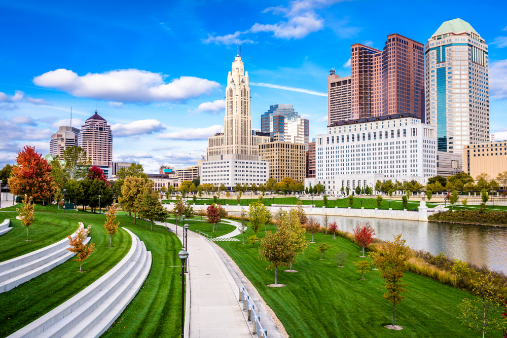 A view of the Columbus Ohio city skyline, a great stop for Ohio road trips. There are several tall buildings that are white, tan, and brick. They are along the river and next to the river is a park with lots of green space and trees. The trees have leaves changing to red and orange.