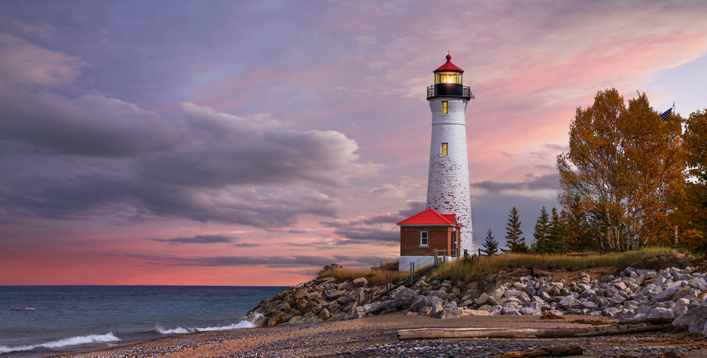 The Crisp Point Lighthouse at the edge of a rocky lake shore. It is a tall white lighthouse with black trim and a red roof. In front of it is a small brick building with a red roof. There are trees with brown leaves near it. It is sunset and the sky is pink and purple