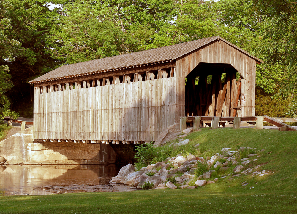 A large wooden covered bridge that crosses a small river. There is a grassy hill next to the road that leads to the bridge and there are rocks along the shore of the small river. The covered bridge is surrounded by trees on the other side, all with green leaves. 