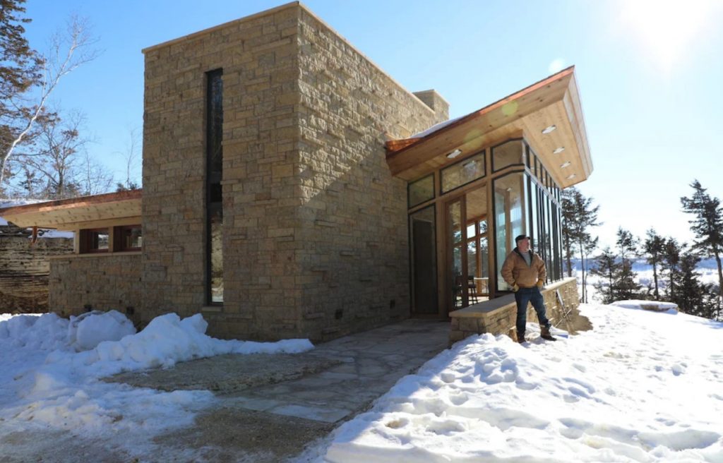 The exterior of a Frank Lloyd Wright inspired cottage on a mountain. The structure is very angular and made of natural stone and wood. To the side there is a large space that is walls of windows. There is snow on the ground and a man standing proudly in front of the cottage.
